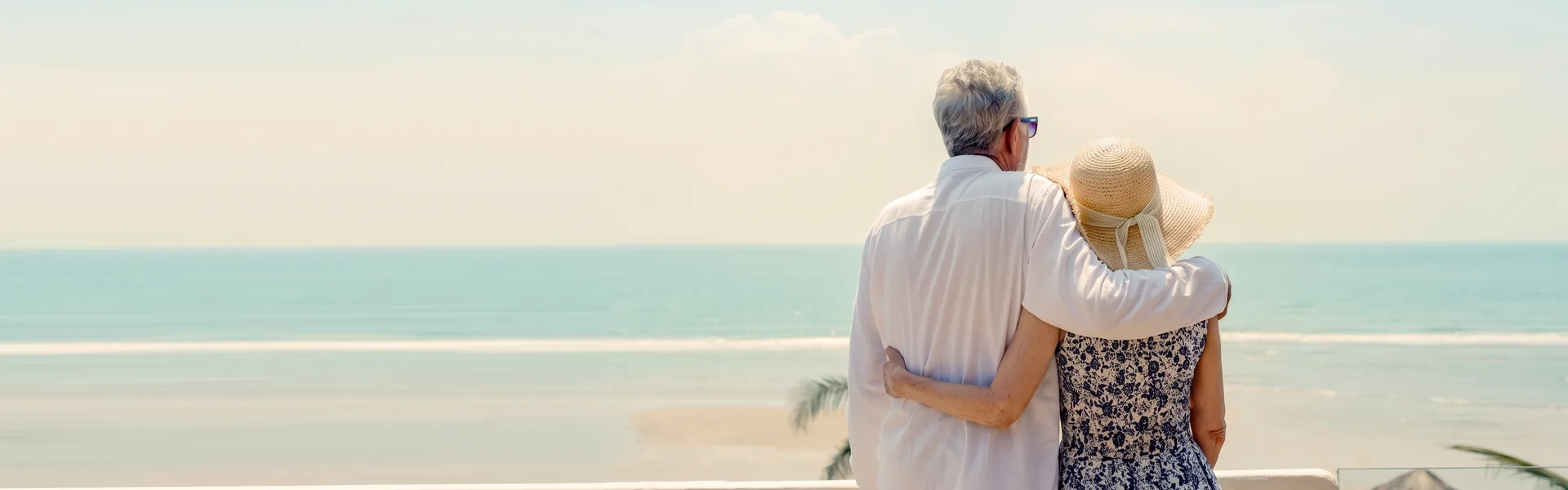 Casal de idosos abraçado, de costas, olhando o mar de uma varanda